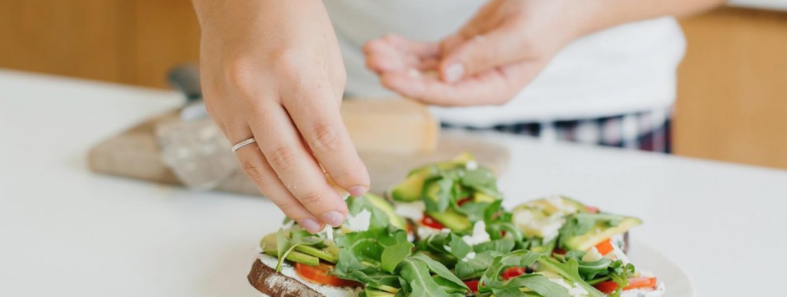 Sandwich made of whole grain bread, avocado, tomato, arugula and cheese. Home cooking concept. Hands putting parmesan on homemade toasts with avocado on modern white kitchen.